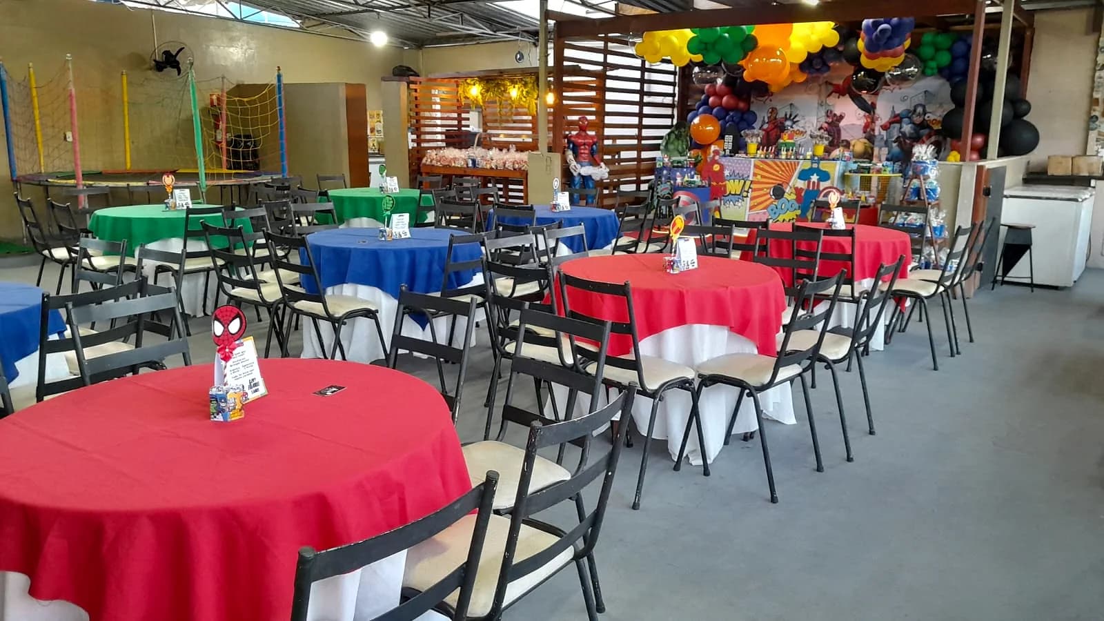 Salão com mesas coloridas e decoração de super-heróis ao fundo